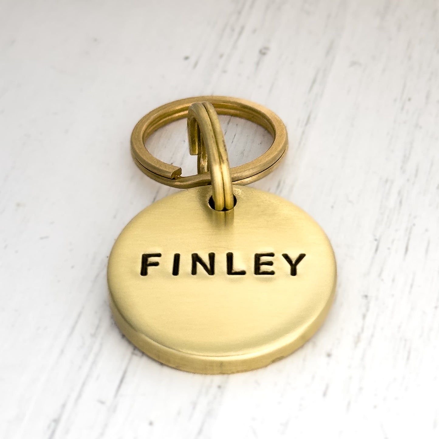 Thick brass dog id tag personalized with the name 'FINLEY' on a light wooden surface.